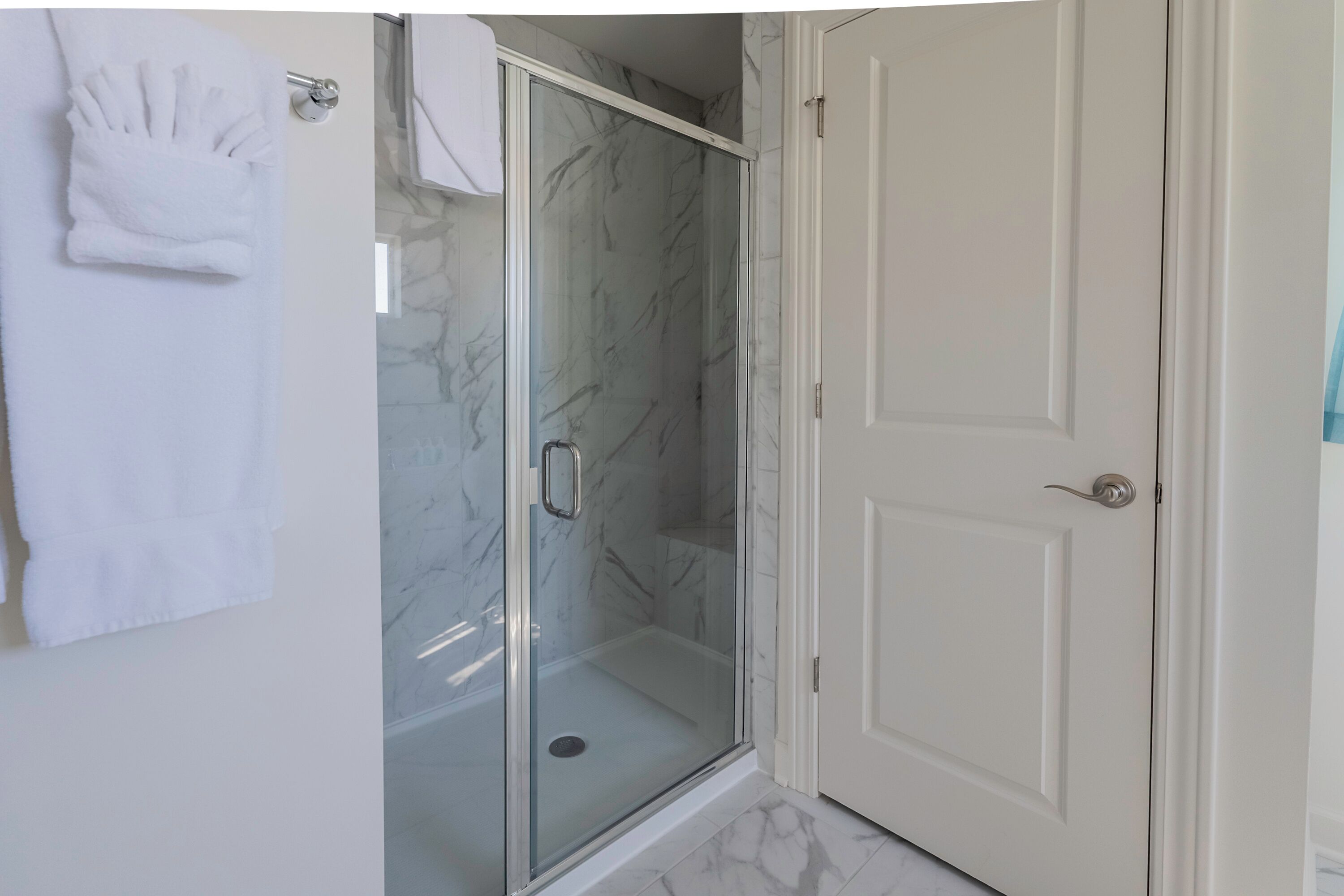 Un baño con cabina de ducha de vidrio, una puerta blanca y un perchero con toallas dobladas.