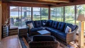 A cozy living room with a large sectional sofa, wicker chairs, and a coffee table. Large windows offer a view of a garden and a body of water.