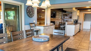 A modern kitchen with a dining area, featuring a large clock, pendant lights, and a rustic brick wall.