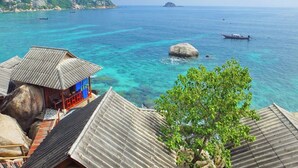 A coastal view with clear turquoise waters, a large rock in the sea, and traditional buildings with corrugated roofs. - Peaceful Seaview Bungalow Koh Tao Snorkeling and Relaxing Paradise Sharkbay (Koh Tao)