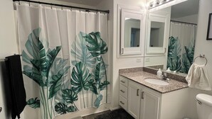 Une salle de bain avec un rideau de douche orné de grandes feuilles vertes, un lavabo blanc avec un plan de travail en granit, un miroir au-dessus du lavabo et des toilettes.