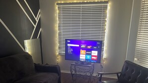 Un salon avec un téléviseur à écran plat fixé au mur, un canapé, une table basse avec des télécommandes et une lampe.