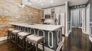 Una cocina con pared de ladrillo, armarios blancos, una isla central con taburetes de bar y un refrigerador de acero inoxidable.