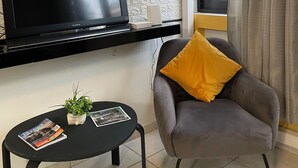Un salon moderne avec un téléviseur à écran plat fixé sur une étagère noire, un fauteuil gris avec un coussin jaune, une petite table ronde avec une plante et des magazines, et une fenêtre avec des rideaux gris.