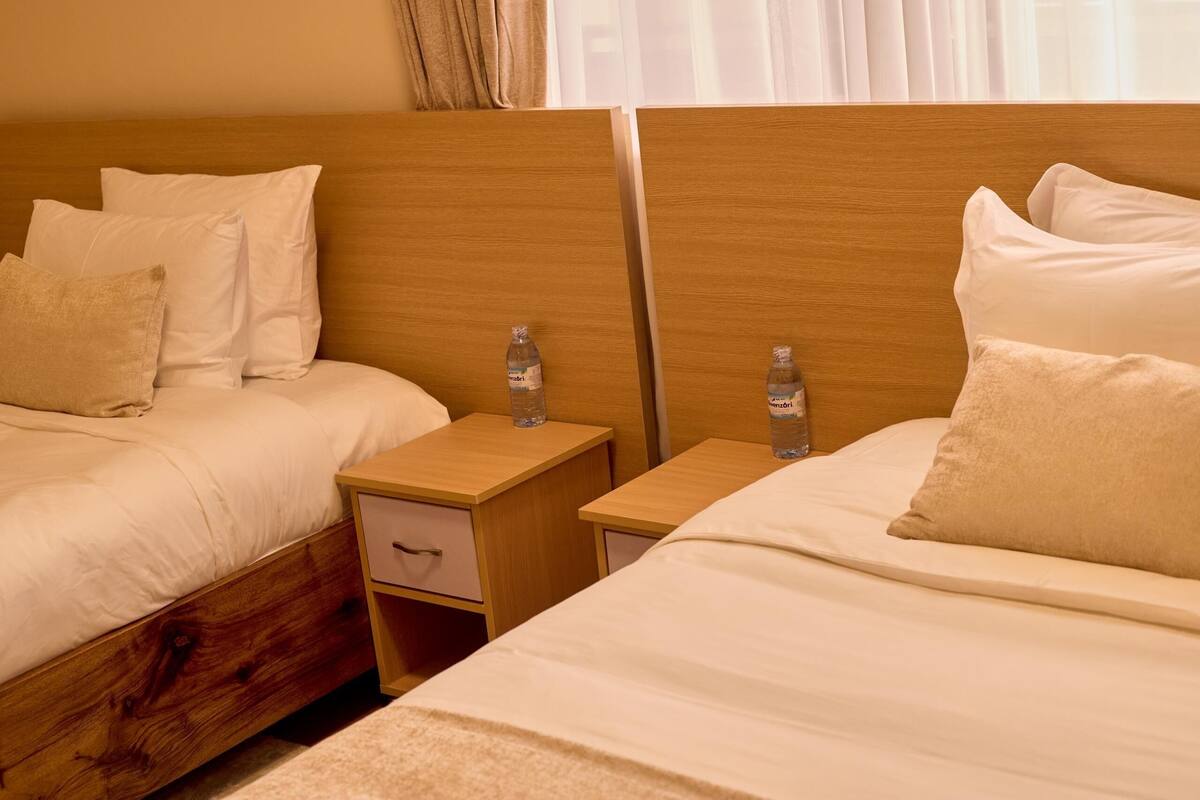 Quarto de hotel com duas camas, cada uma com roupa de cama e almofadas brancas. Há mesas de cabeceira de madeira com garrafas de água.