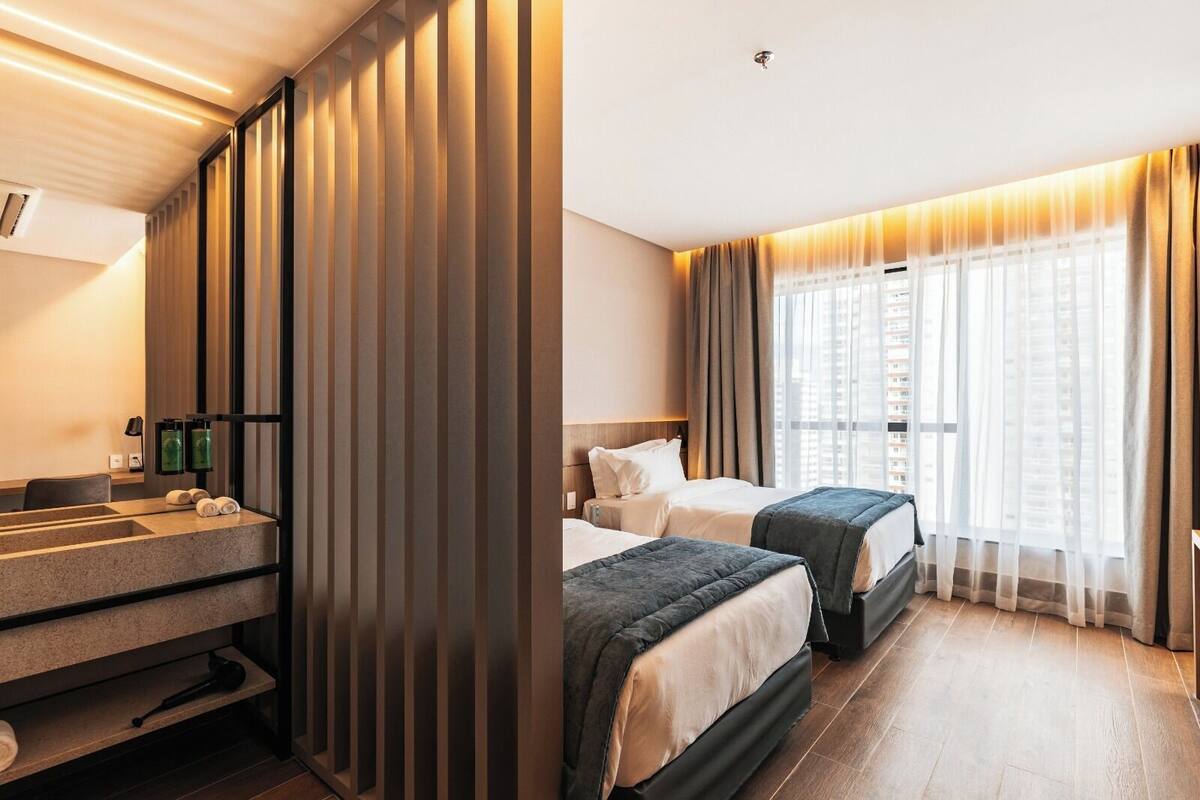 Ein Hotelzimmer mit großem Fenster, einem Bett, einem Nachttisch und einem Badebereich mit Waschbecken und Handtuchhalter.