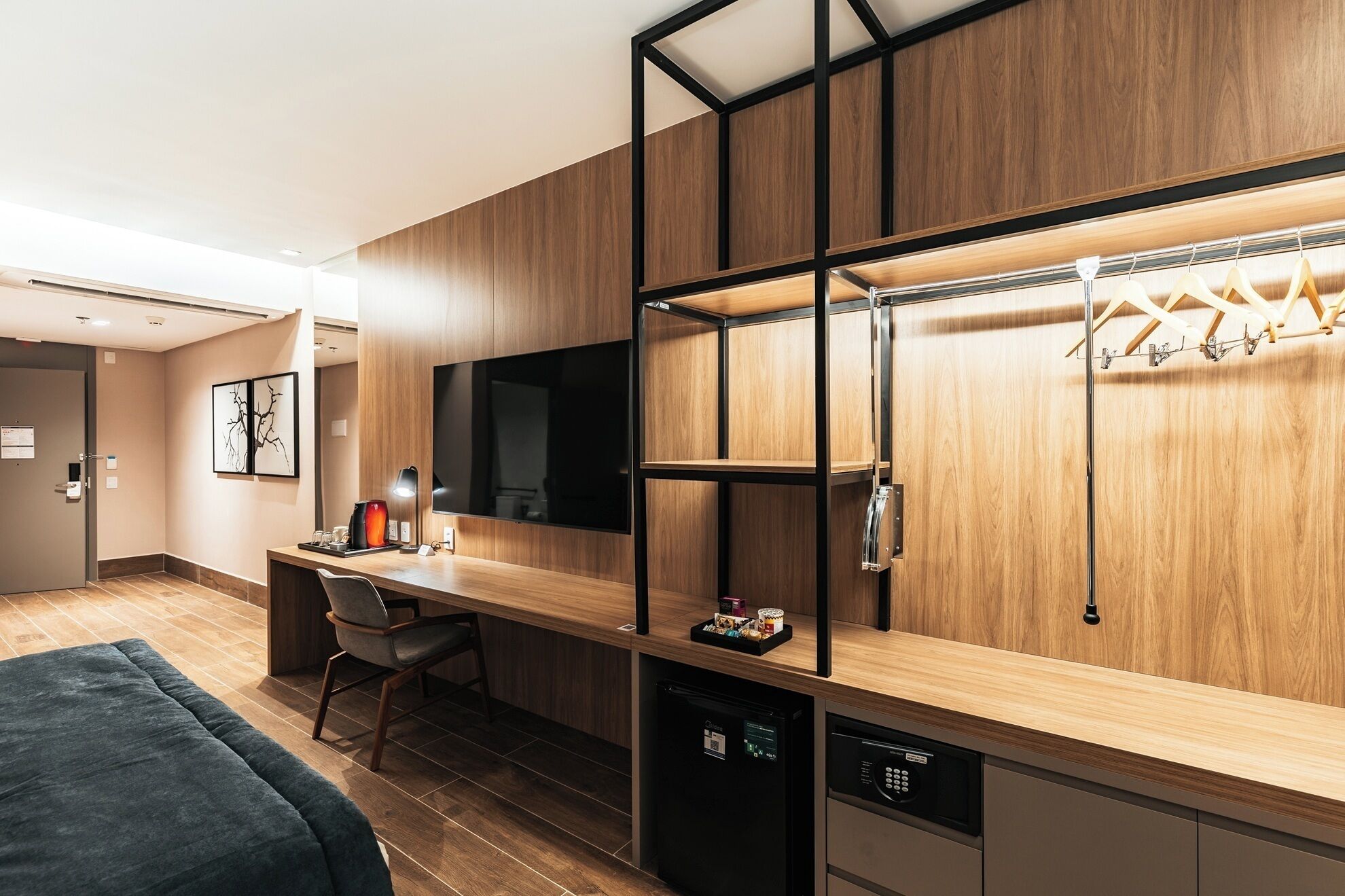 A modern hotel room with a wooden desk, a flat-screen TV, a built-in wardrobe, and a mini-fridge.