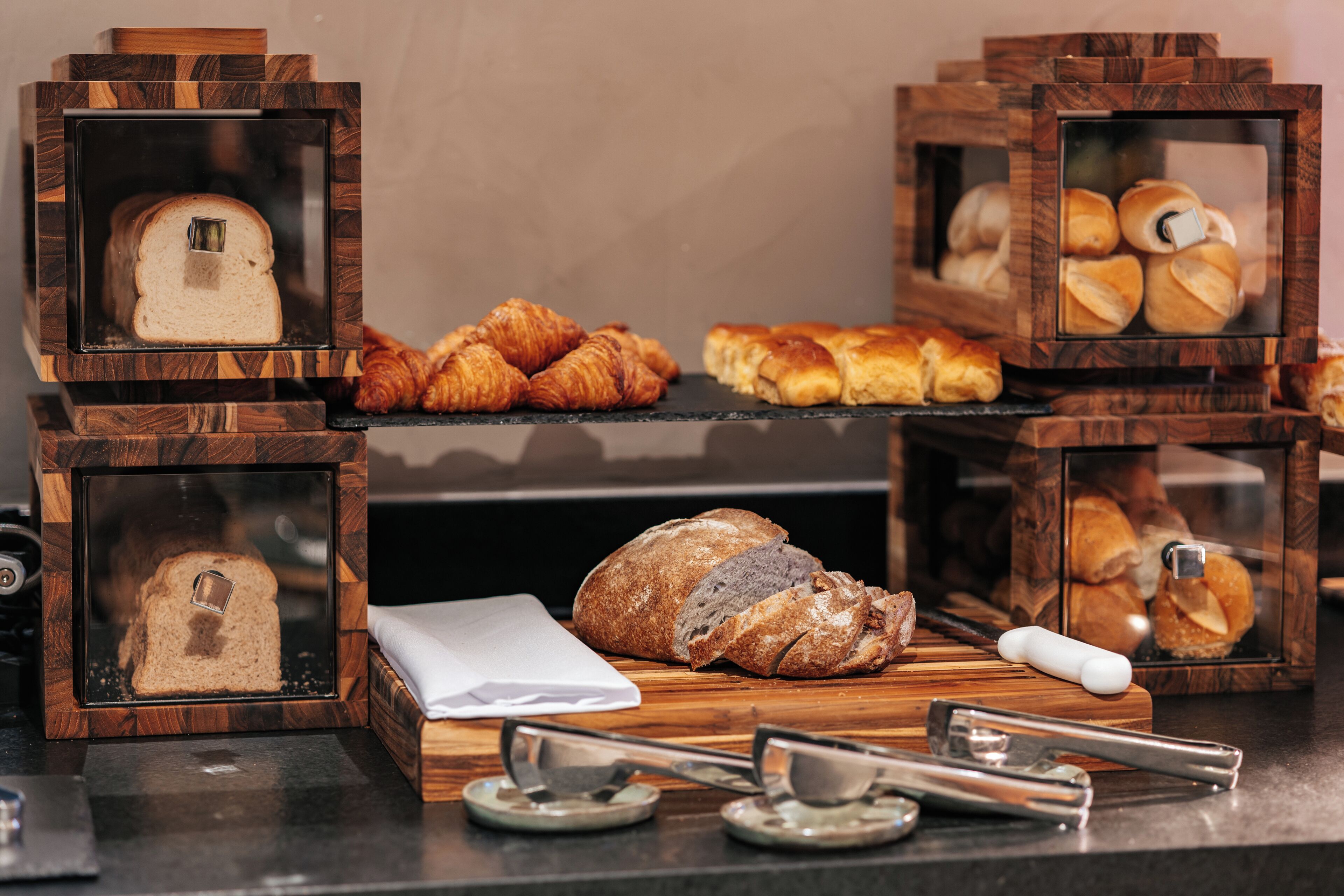 A display of various breads and pastries in wooden cases, with a loaf of bread and a baguette on a wooden board.