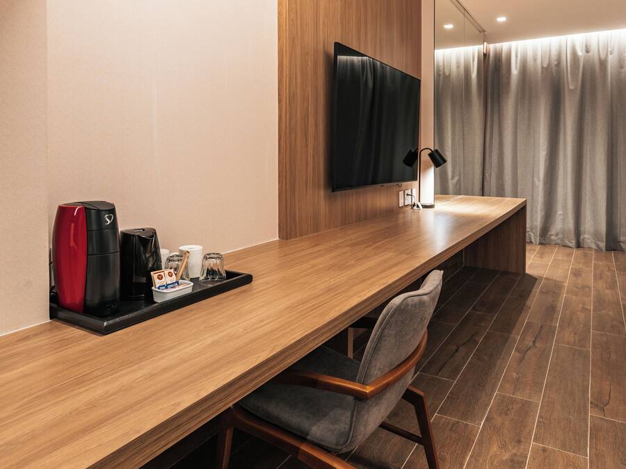 Una habitación de hotel moderna con un escritorio de madera alargado, un televisor de pantalla plana montado en la pared, una cafetera y una silla.