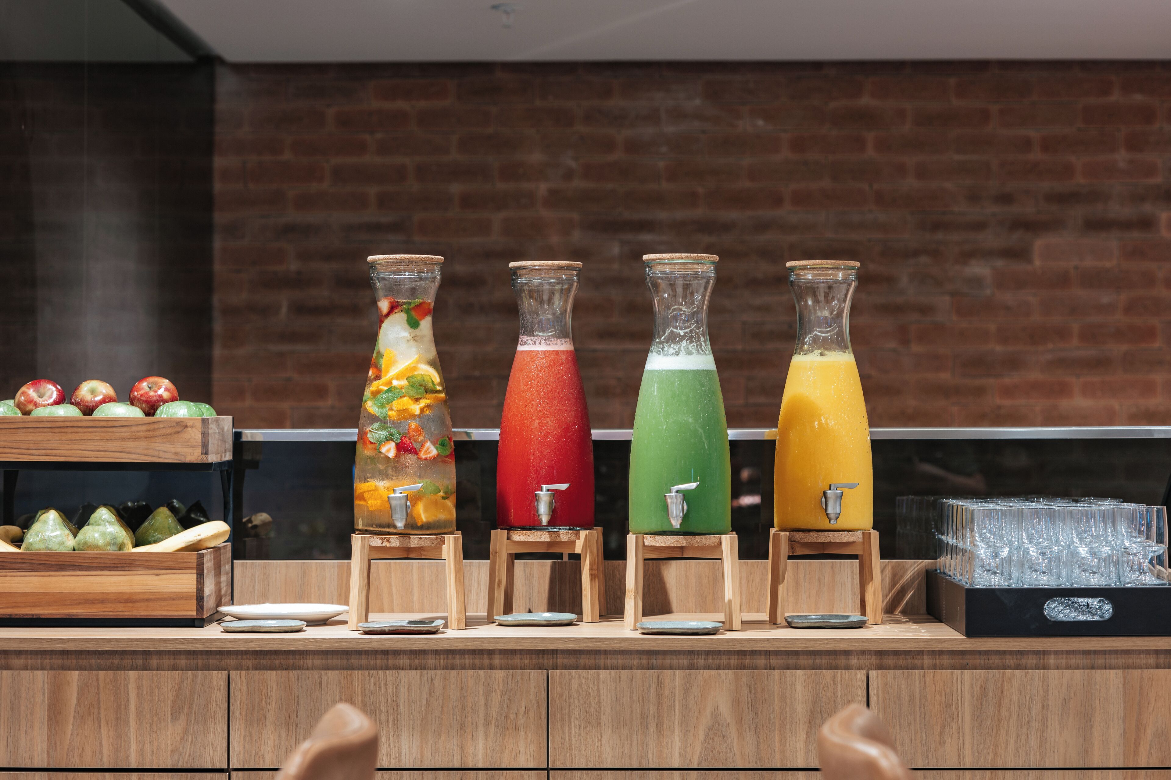 En juicebar med fire dispensere, hver med en forskelligfarvet drik, samt en trædisk med tallerkener og et frugtdisplay.