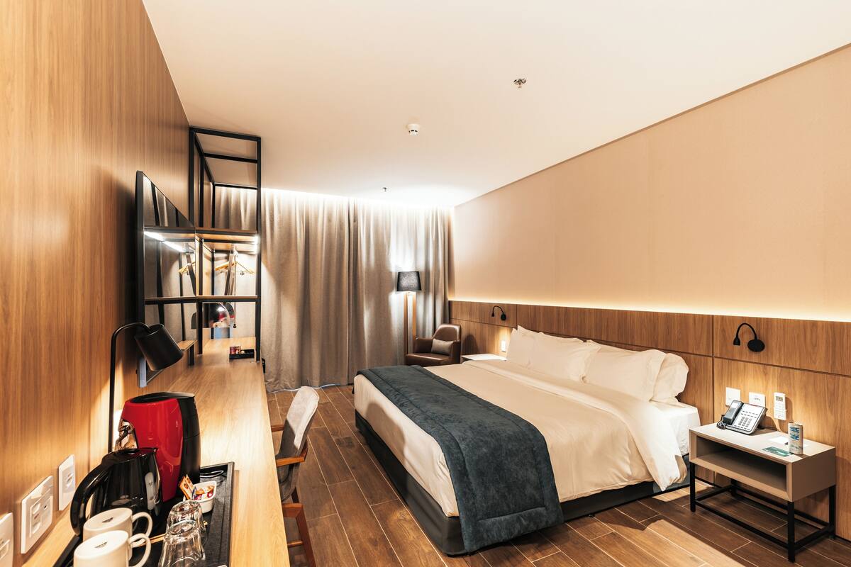 Ein modernes Hotelzimmer mit einem ordentlich bezogenen Bett, einem Nachttisch mit Lampe, einem kleinen Schreibtisch mit Telefon und einer Sitzecke mit Sessel und Sofa.