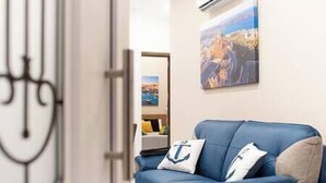 A room with a blue sofa, a wall-mounted air conditioner, and a framed picture of a coastal scene.