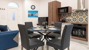 A modern living room with a round glass table, four black chairs, a blue sofa, and a kitchen area with a microwave and patterned tiles.