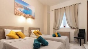 A hotel room with two beds, each with a headboard, and a painting on the wall. There is a window with curtains, a chair, and a small table.
