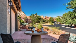 A patio with two wicker chairs, a round table with a vase and a mug, and a mountain view in the background. - Stylish West Sedona Oasis. Hot Tub, E-BIKES & Kids Play Area (Sedona)