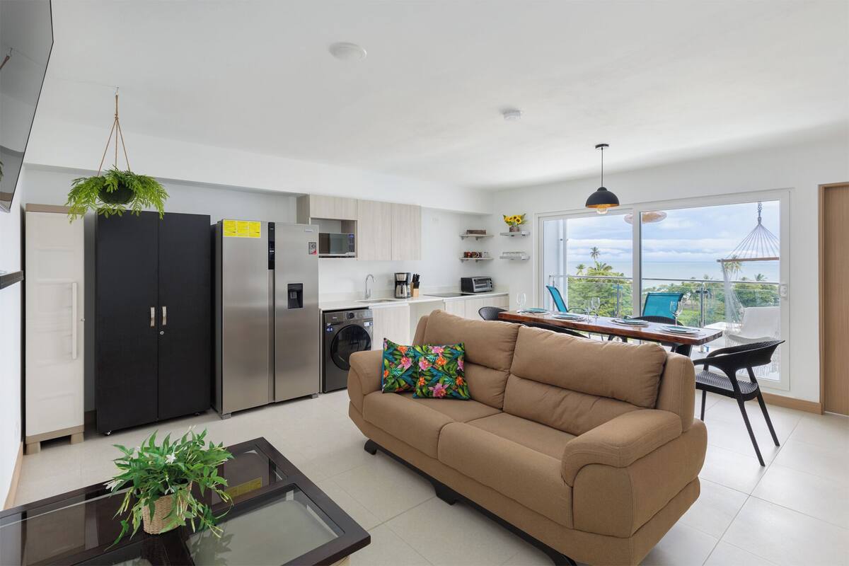 A modern kitchen with a refrigerator, microwave, and washing machine. There's a sofa with a floral pillow, a dining table with chairs, and a balcony with a view.