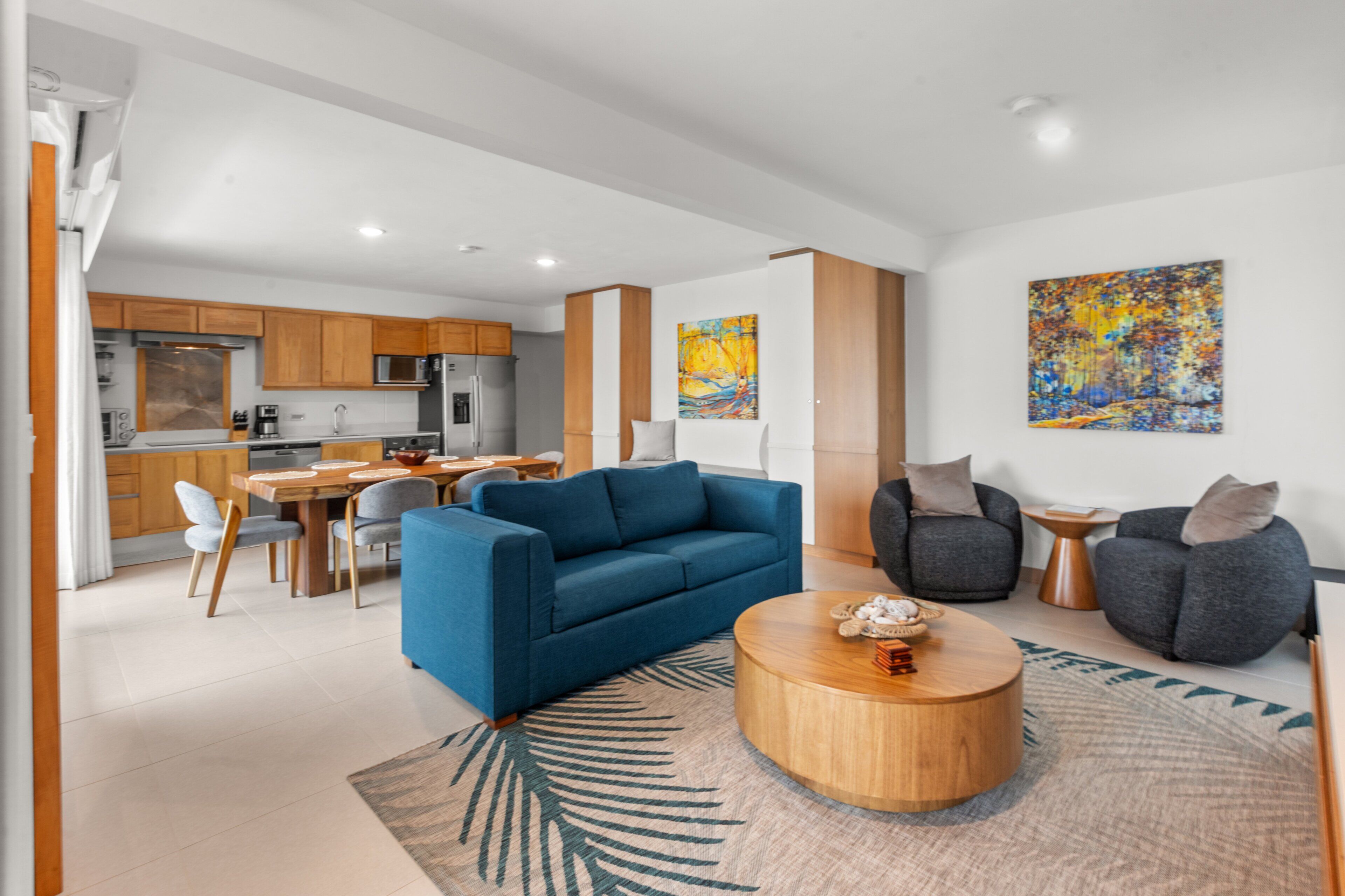 Una sala de estar moderna con un sofá azul, sillones grises, una mesa de centro de madera y un comedor con mesa y sillas de madera.