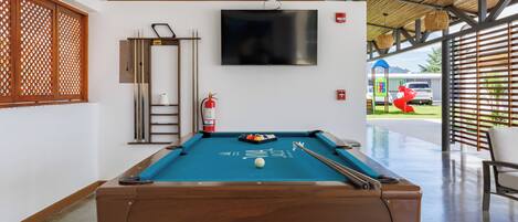 Een biljarttafel met een blauwe viltbekleding, een televisie erboven en een brandblusser aan de muur.