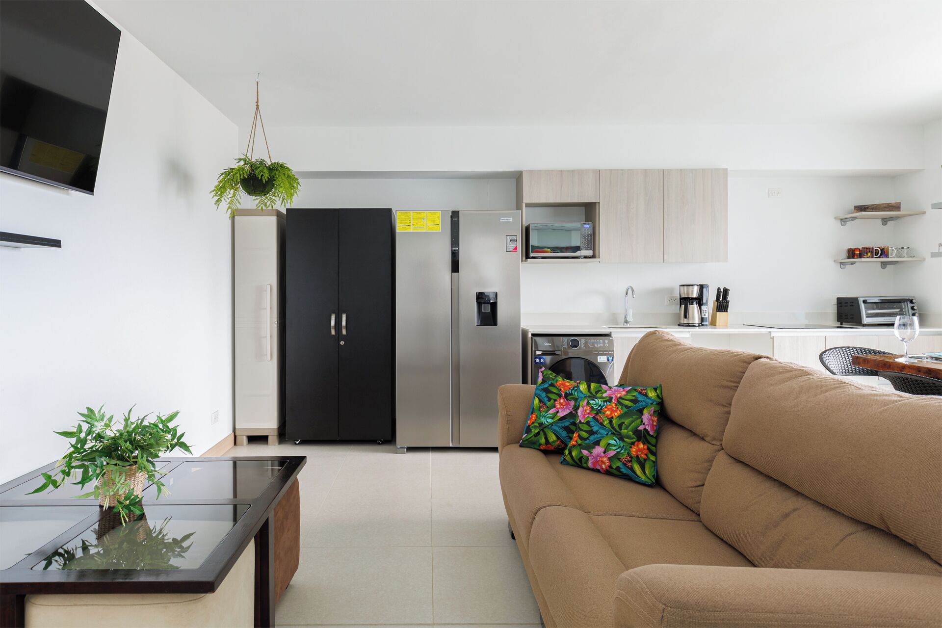 A modern kitchen with a refrigerator, microwave, and coffee maker. There is a sofa with a floral pillow and a small table with a plant.