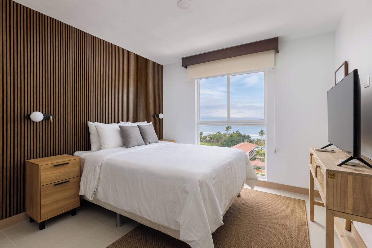 A modern bedroom with a large bed, wooden panel wall, a TV on a wooden stand, and a view of the outdoors.