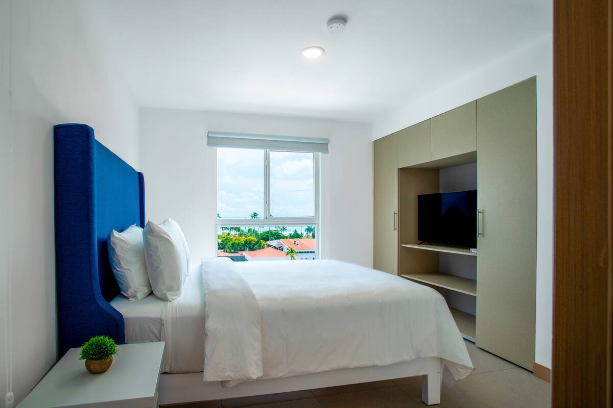 A modern bedroom with a bed, a television, a wardrobe, and a window with a view of buildings and greenery.