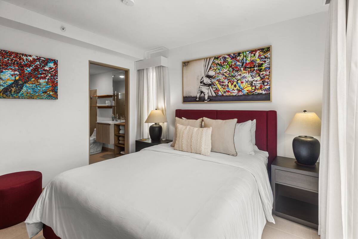 A hotel room with a bed, two bedside tables with lamps, a red headboard, and two paintings on the walls.