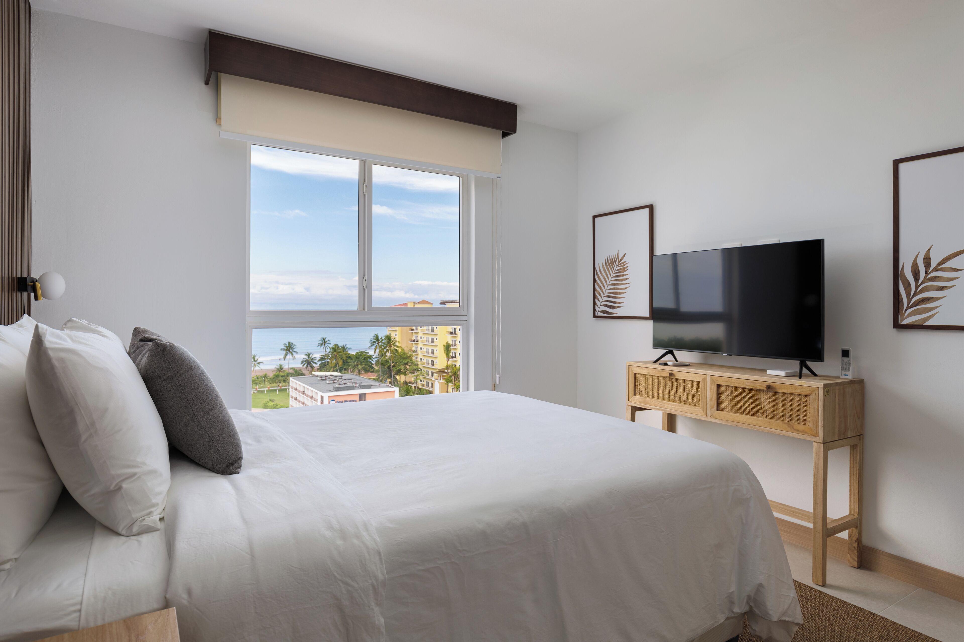 A hotel room with a large bed, a TV, a wooden console table, and a window with a view of palm trees and buildings.
