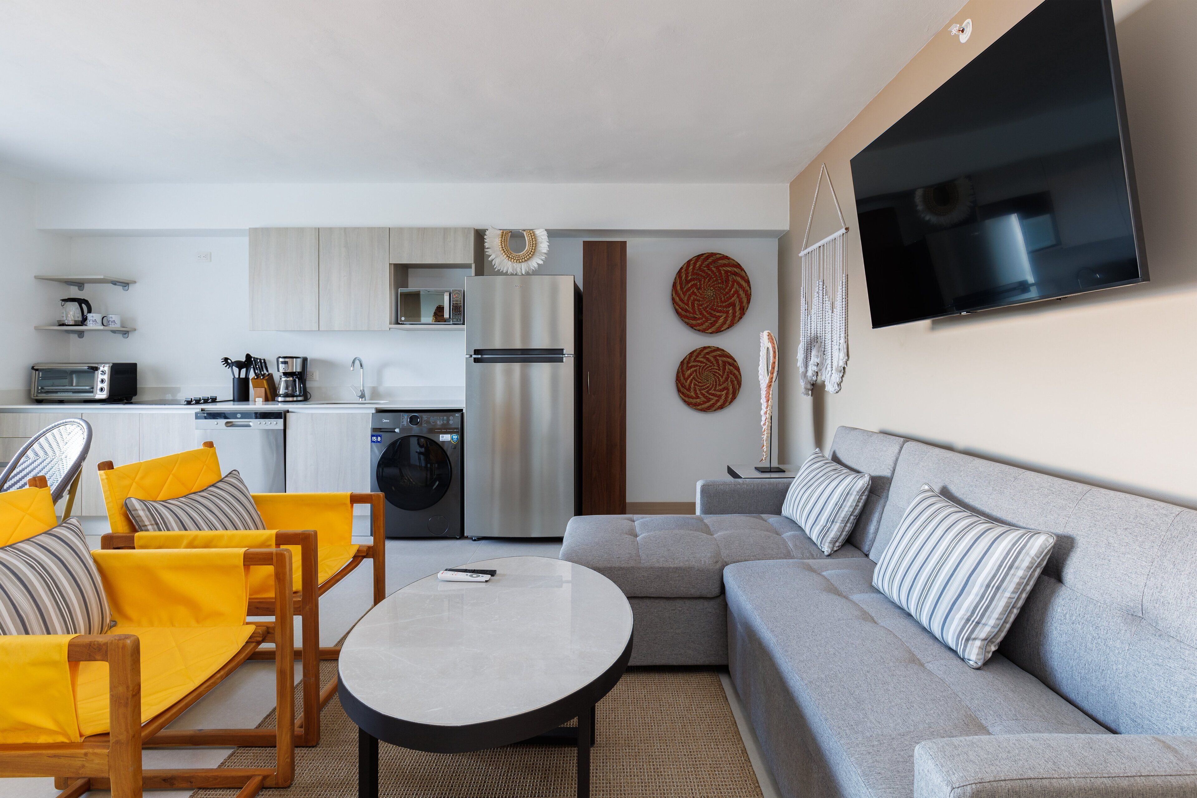 Ein modernes Wohnzimmer mit einem grauen Sofa, gelben Sesseln, einem runden Couchtisch und einer Küchenzeile im Hintergrund.
