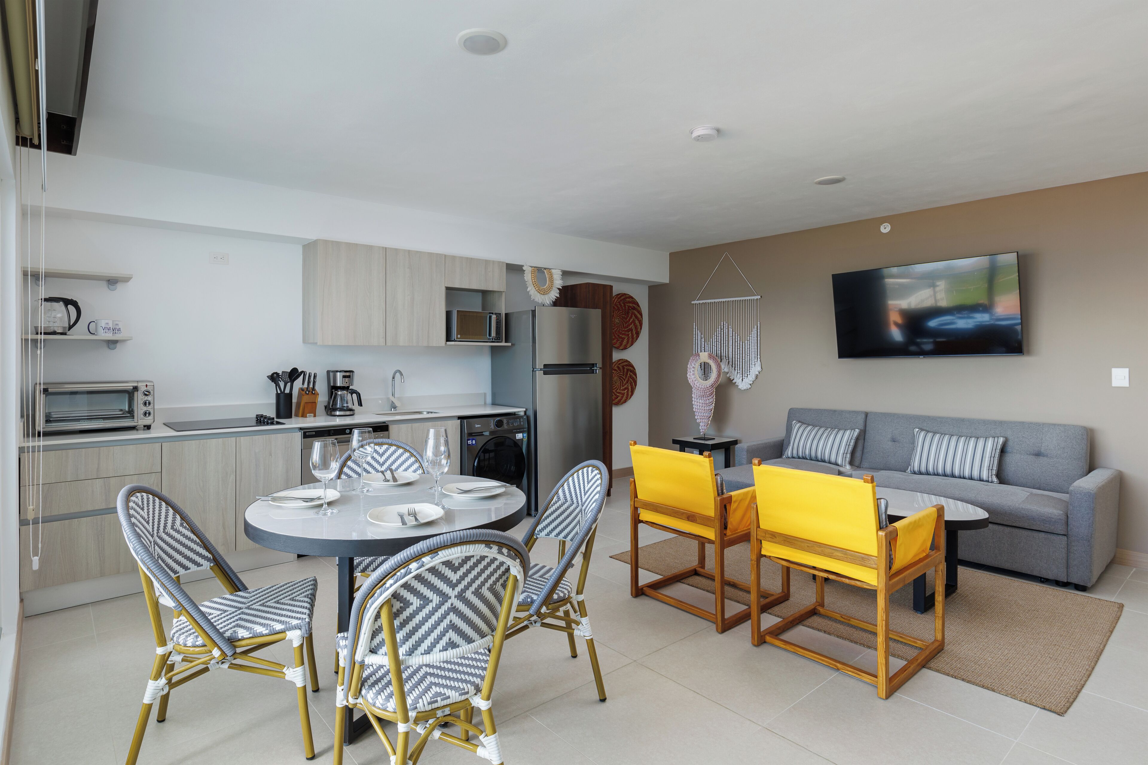 A modern kitchen with built-in appliances, a dining area with wicker chairs and a yellow table, and a grey sofa with striped cushions.