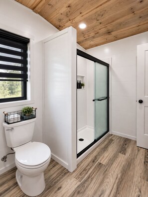 A modern bathroom with a toilet, a shower with a glass door, and a window with blinds. - Peaceful Pastures (Fredericksburg)
