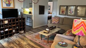 A living room with a television, a coffee table, a sofa, and a lamp. There is a kitchen in the background.