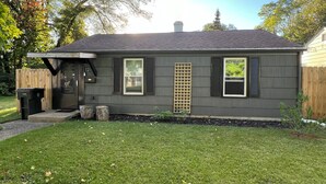 A single-story house with a dark gray exterior, a front door, two windows with shutters, a small porch area, and a wooden fence in the background.