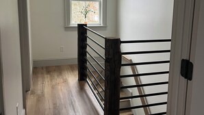 A narrow hallway with wooden flooring, a window with a view of trees, and a staircase with a wooden railing.