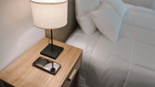 A neatly made bed with white linens, a bedside lamp, and a smartphone on a wooden bedside table.