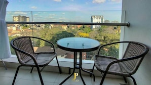 Ein Balkon mit einem runden Tisch und zwei Korbstühlen, mit Blick auf eine Stadtlandschaft mit Gebäuden und Bäumen.