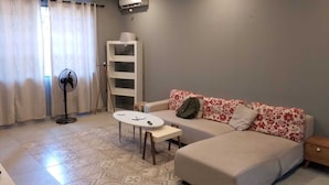 Une petite pièce, bien rangée, avec un lit, un canapé, une petite table, un ventilateur et une étagère.