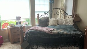 Um quarto com cama, mesas de cabeceira, janela com cortinas e vista para o verde lá fora.