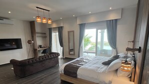 A modern bedroom with a large bed, a dark brown sofa, a TV, and a large window with curtains. - La Parenthèse (Mbodiène)