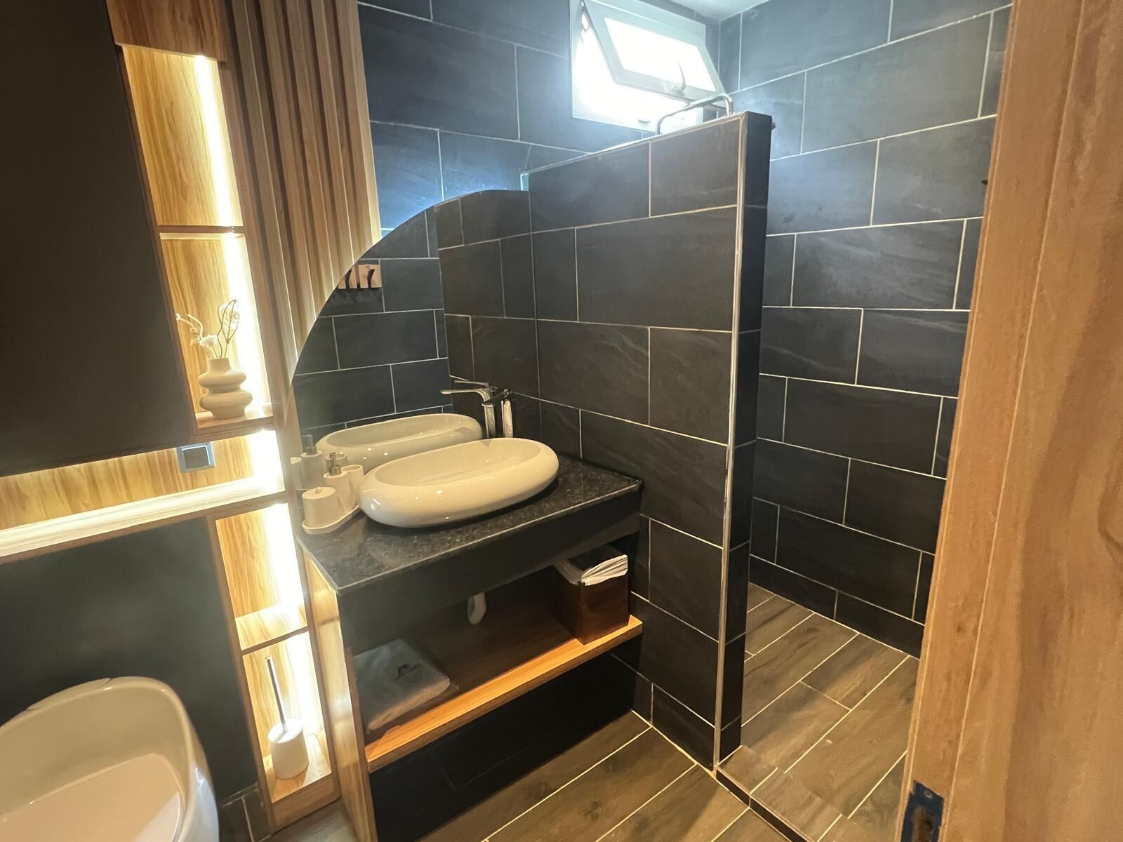 A modern bathroom with dark tiles, a white sink, and a wooden vanity.