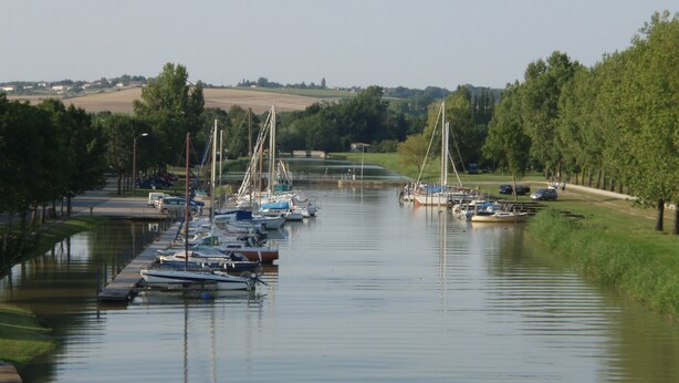 A calm river with boats docked along the banks, surrounded by greenery and a distant bridge.