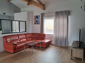 A living room with a red leather sofa, a wooden coffee table, a TV on a wooden stand, and a window with curtains.