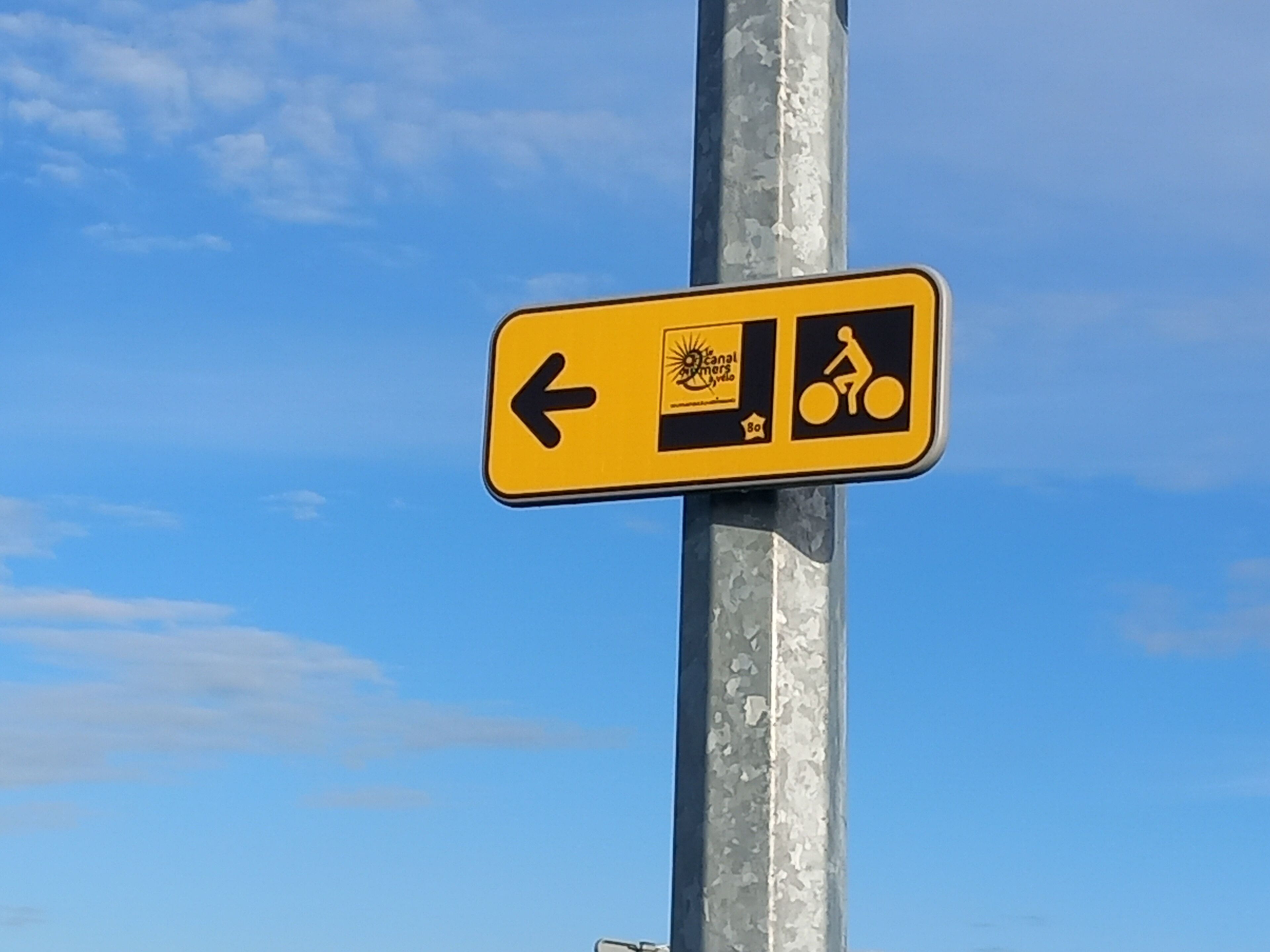 Un panneau de signalisation indiquant la direction, avec un symbole de vélo et un logo représentant un soleil.