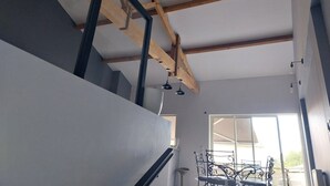 A staircase with wooden beams and a glass railing, leading to a room with a large window and a dining area with metal chairs.