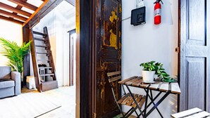 Un interior rústico con una puerta de madera, una pequeña mesa con una planta en maceta, un extintor y una escalera que lleva a otra habitación.