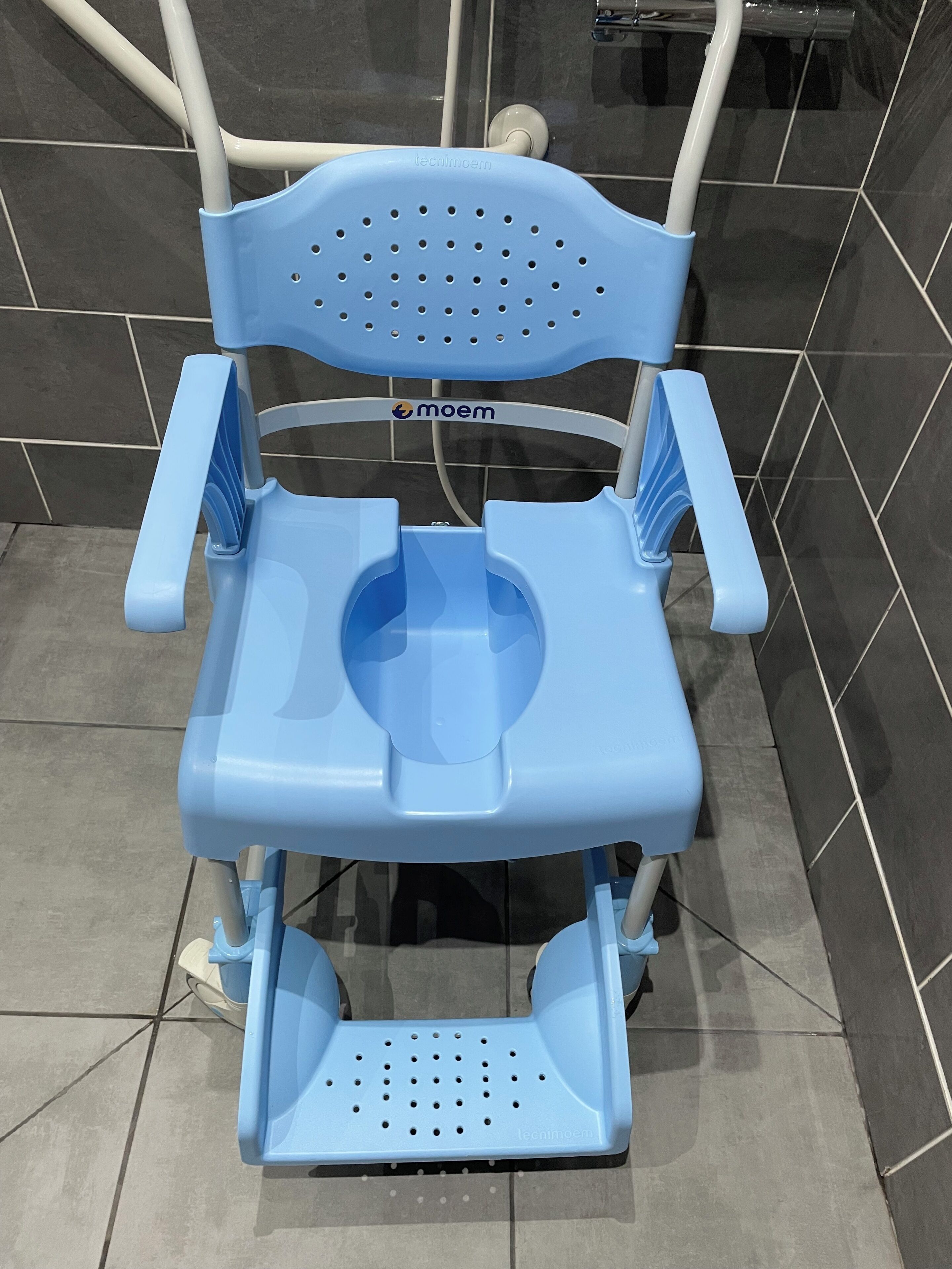 Un baño químico azul portátil con asiento perforado y reposapiés, sobre un suelo de baldosas.