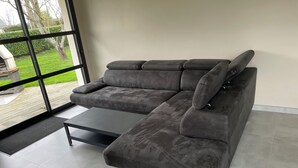 En moderne stue med en grå hjørnesofa, et lille sofabord og et stort vindue med udsigt til en have.
