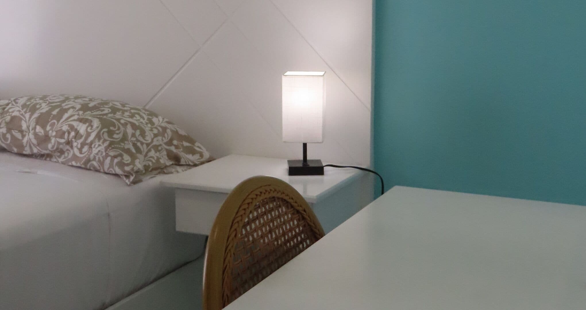 Una habitación pequeña y ordenada con una cama, una mesita de noche con lámpara, una silla y una pared color verde azulado.