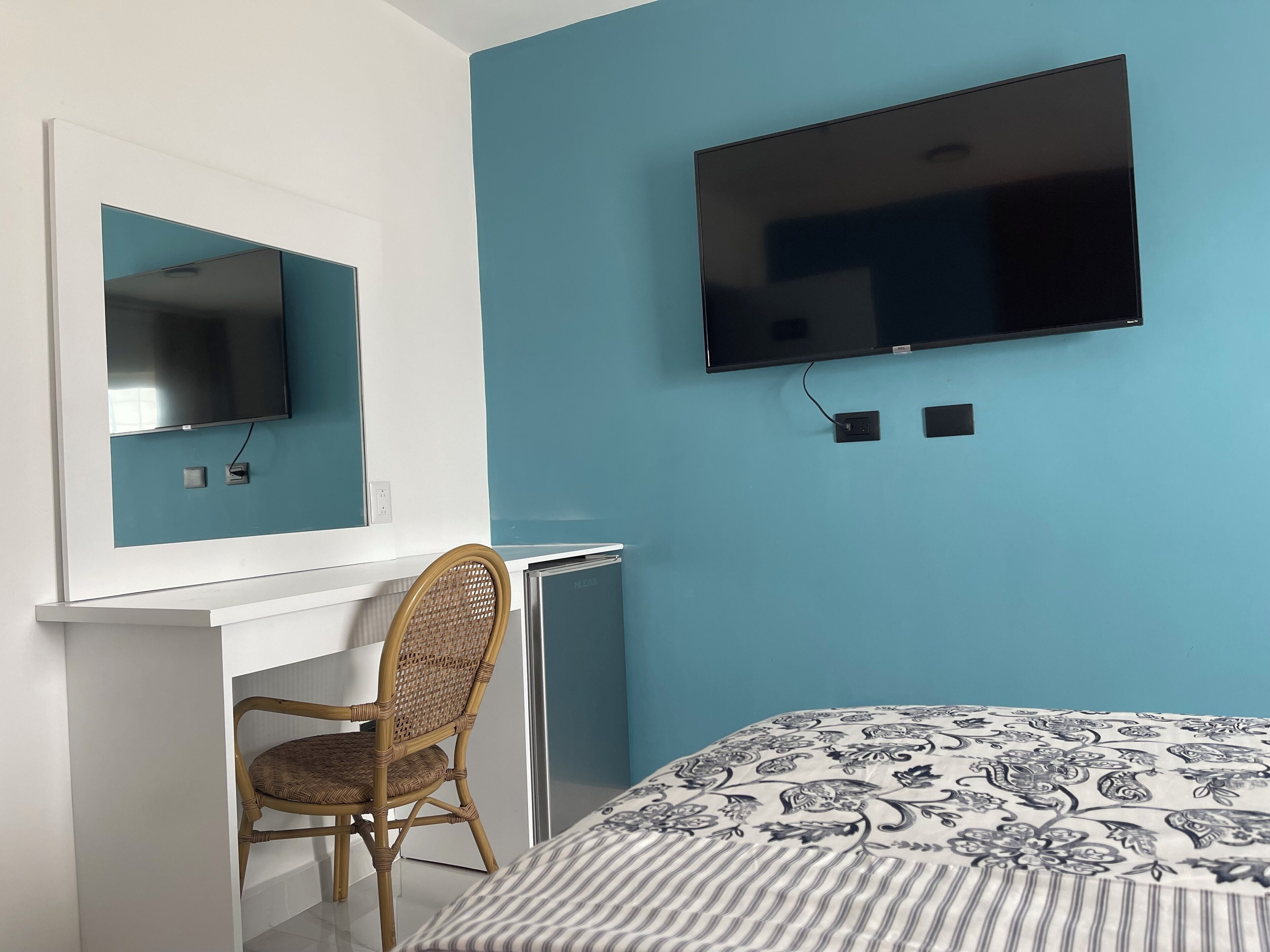 A bedroom with a bed, a TV mounted on the wall, a desk with a chair, and a window.