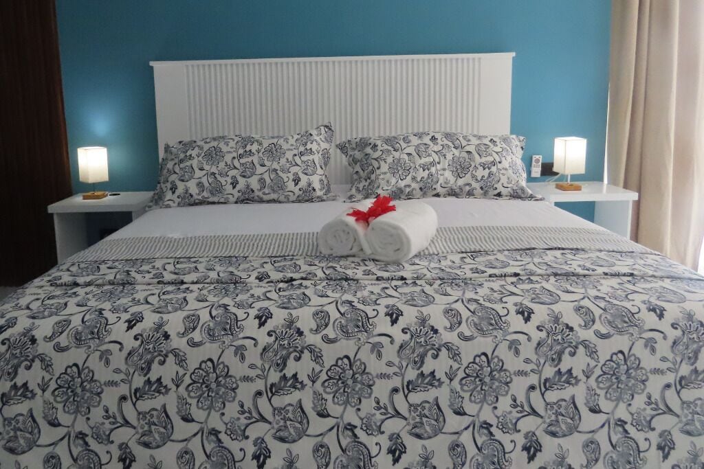 Una cama bien hecha con ropa de cama de estampado floral, dos almohadas con un diseño similar y un par de toallas con un lazo rojo.