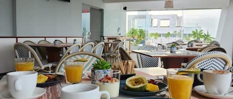 Una mesa de desayuno con diversos platos, bebidas y mobiliario de mimbre en una habitación con ventilador de techo y ventanas amplias.